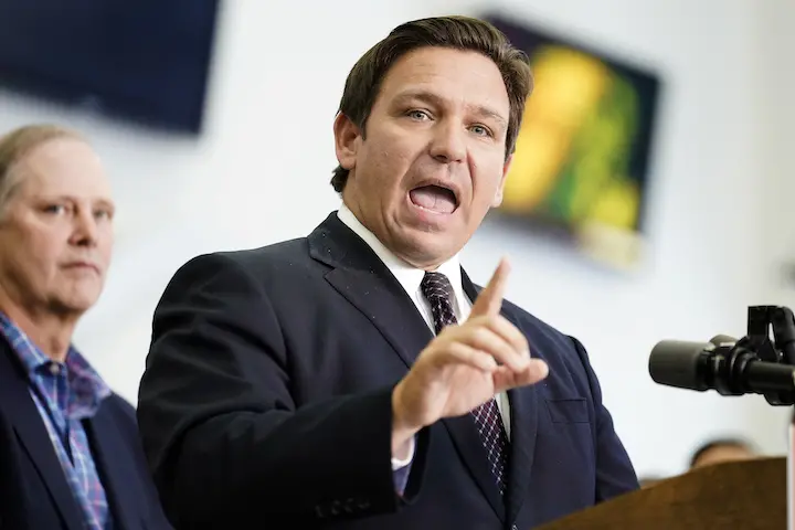 A man in a dark suit and tie speaks emphatically at a podium, raising his index finger, possibly addressing topics like the DeSantis Stop WOKE Act. Another man in a plaid shirt stands in the background, slightly out of focus.