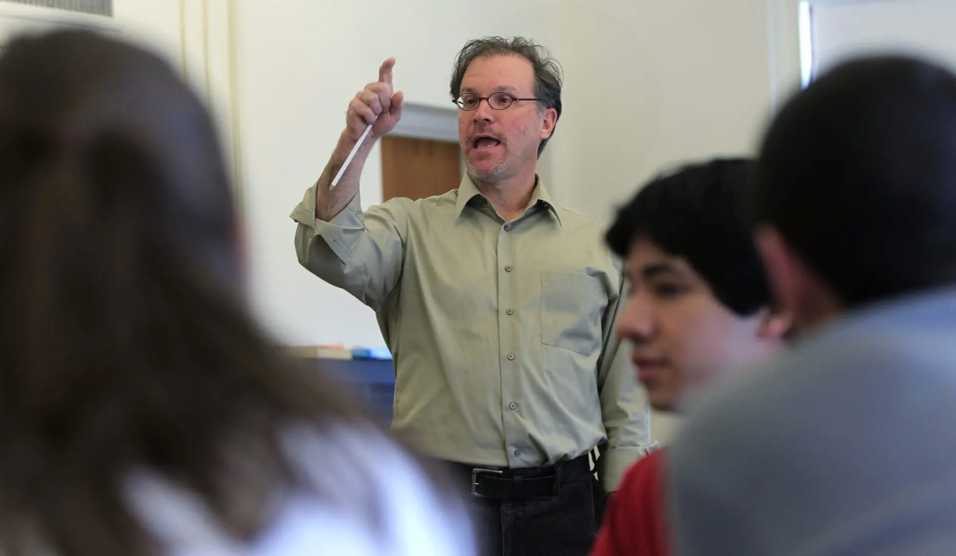 A teacher wearing glasses and a light green shirt gestures while speaking to students in a classroom, highlighting the challenges posed by educational gag orders. The focus is on the teacher, with students blurred in the foreground.