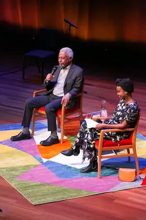 Two people sit on stage in colorful chairs, engaging in conversation. The man on the left holds a microphone, while the woman on the right looks at her notes. A vibrant, geometric rug covers the floor beneath them.
