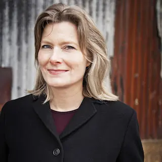 A woman with shoulder-length blonde hair wearing a black coat stands in front of a weathered, corrugated metal wall, smiling slightly at the camera, evoking the introspective style of Jennifer Egan’s pen.