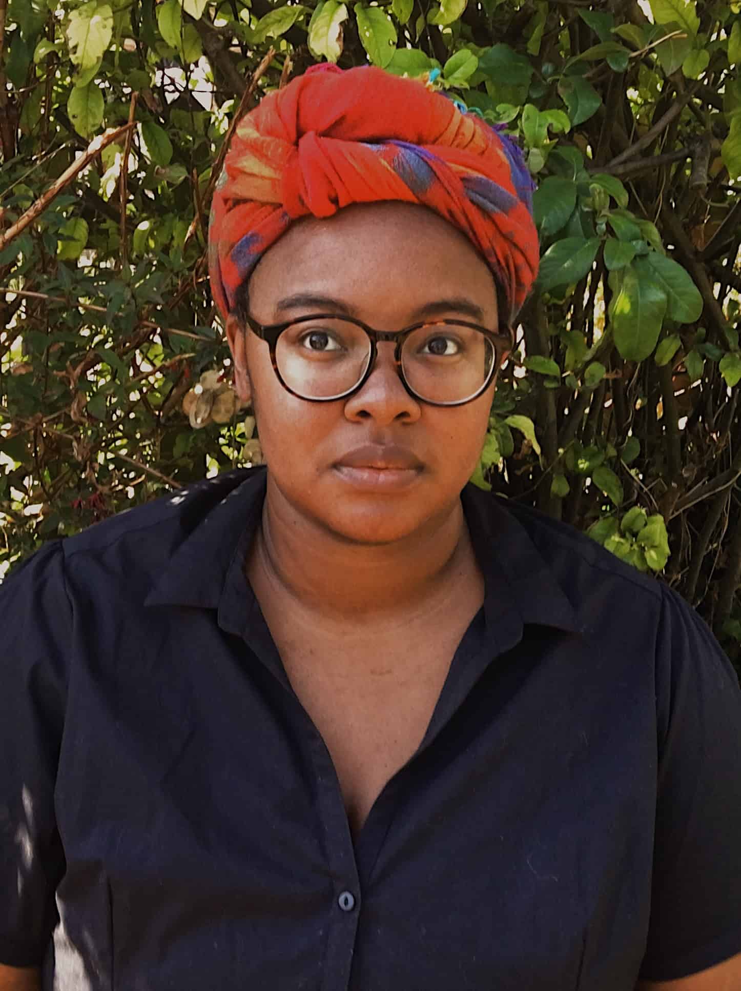 A person wearing glasses, a bright red and orange headwrap, and a dark shirt stands in front of leafy green bushes, looking directly at the camera with a neutral expression, reminiscent of a Nicole Chung interview portrait.