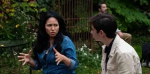 Two people sit outside in a garden, engaged in a serious conversation. Raquel Almazán gestures with her hands while talking, as a third person sits blurred in the background among greenery.