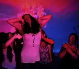 A group of women stands in colorful lighting; Raquel Almazán is at the center, posing with arms raised and eyes closed, while others gently touch and support her, creating a scene of solidarity or empowerment.