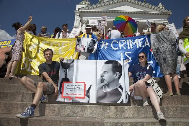 A group of protesters sit and stand on steps, holding banners including a large photo of Oleg Sentsov, signs reading #CRIMEA IS and FreeSentsov, and a yellow-blue flag, in front of a white building.