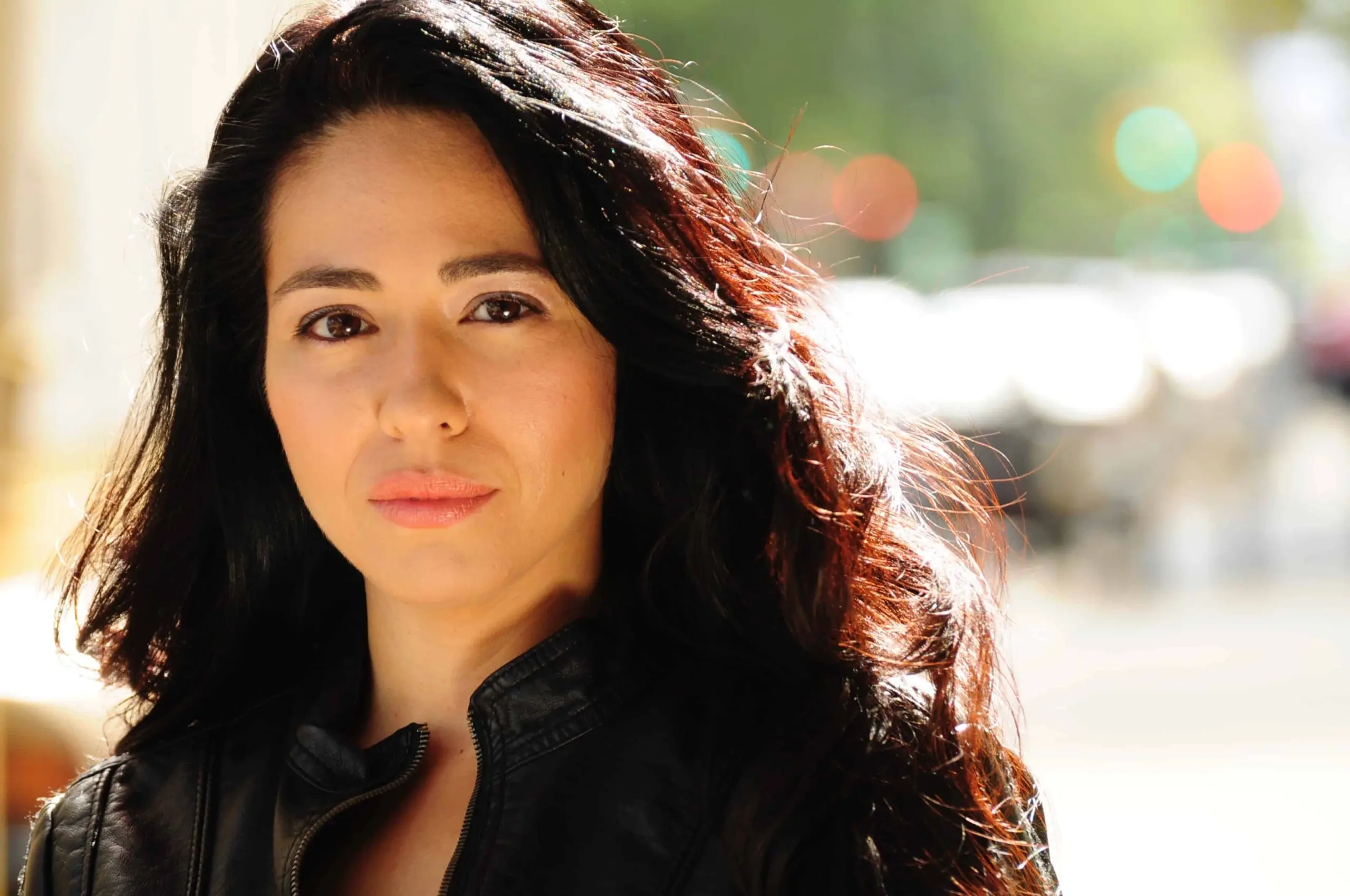 Raquel Almazán, with long dark hair and a black jacket, stands outdoors in sunlight, looking directly at the camera with a neutral expression. The blurred background features green and orange tones.