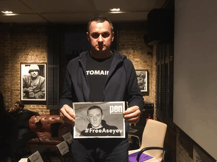 A man stands indoors holding a sign with a black-and-white photo and the text #FreeAseyev and pen. The background features brick walls, framed photos, and some people sitting on chairs.