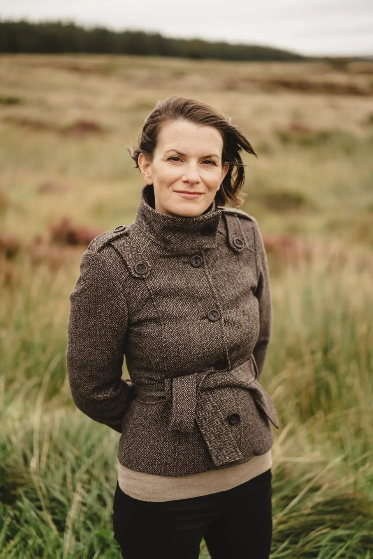 A woman in a brown belted coat stands in a grassy field, her hair blowing in the wind and a slight smile on her face, as if pausing mid-thought during a Michelle Gallen interview. Tall grass and distant trees fill the open landscape behind her.