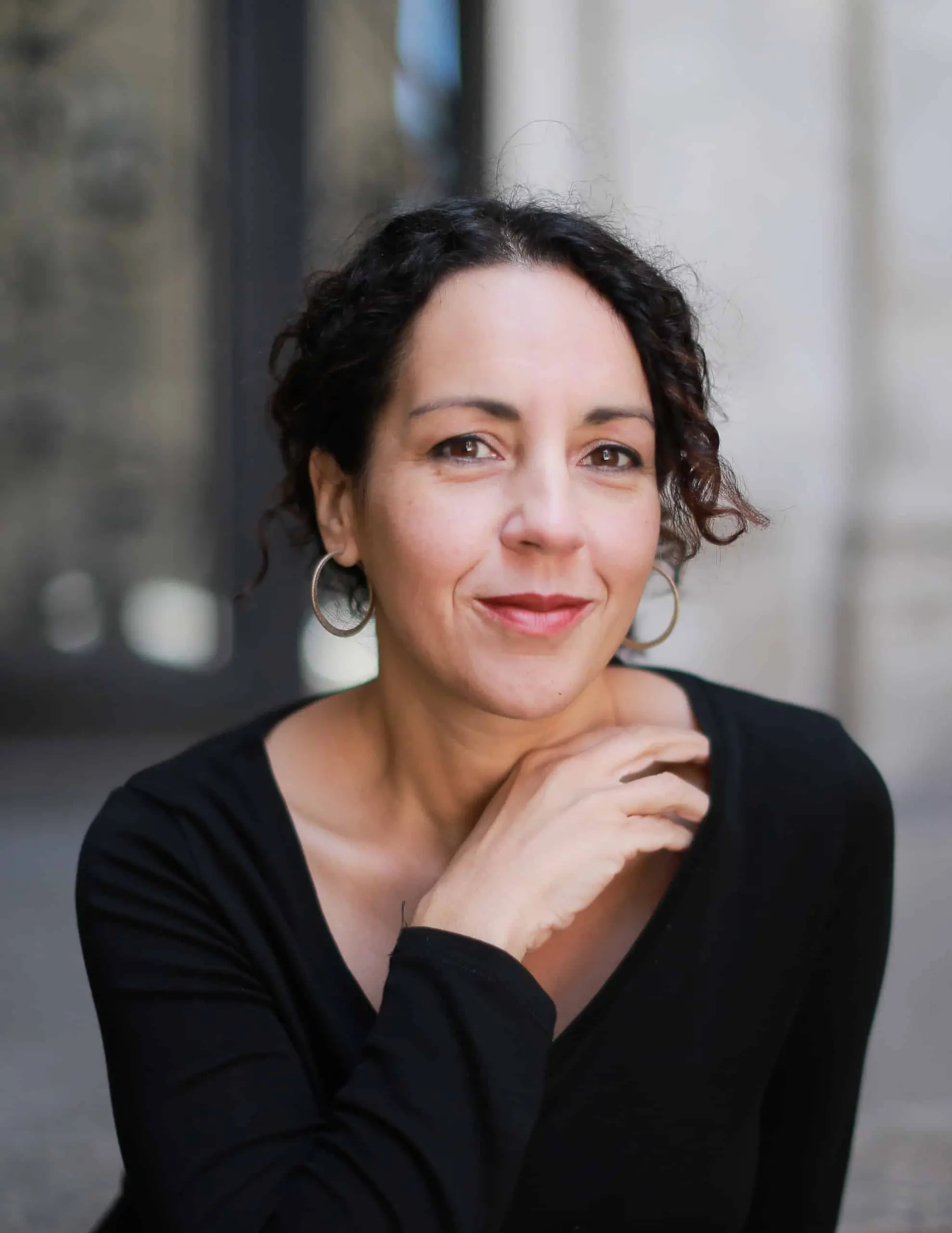 A woman with curly dark hair, wearing a black top and hoop earrings, smiles softly at the camera while resting her hand near her chin, reminiscent of Azar Nafisi holding a pen. The background is softly blurred.