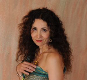 A woman with long, curly dark hair poses against a textured, peach-colored background. Reminiscent of a Sholeh Wolpe interview, she wears a strapless green and gold dress, gold jewelry, and looks at the camera with a gentle smile.