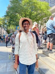 A smiling person wearing glasses, a wide-brimmed hat, a light shirt, and a backpack stands outdoors near steps, with a group of people and trees in the background on a sunny day.