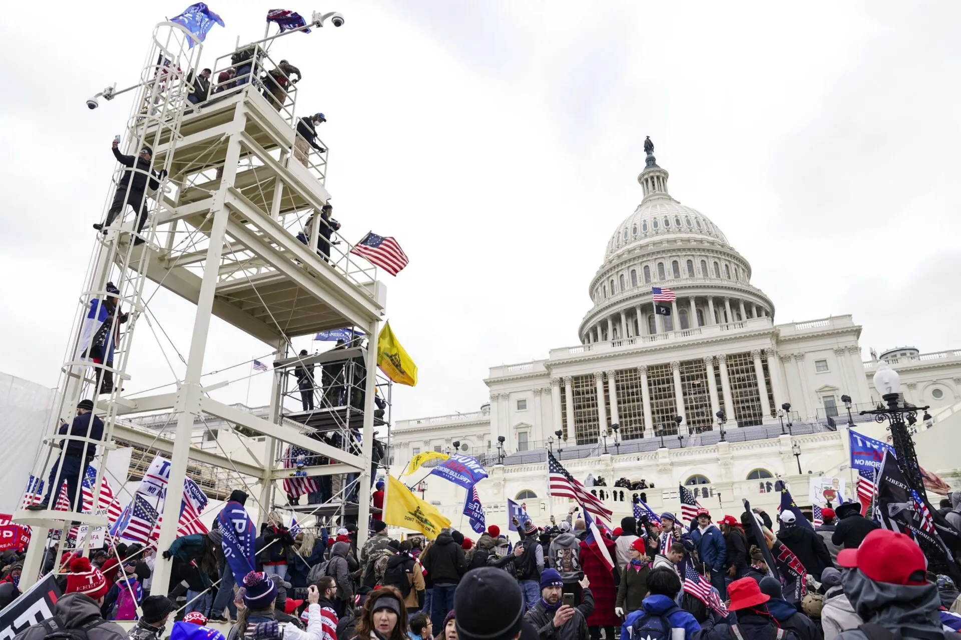 A large crowd of people with American flags and banners gathers outside the U.S. Capitol building; some stand on scaffolding, and the Capitol dome is visible in the background under a cloudy sky.