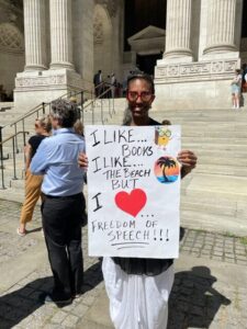 A person stands outside a large building holding a sign that reads, I like… books, I like… the beach, but I love… freedom of speech!!! with cartoon drawings of a book, palm tree, and heart.