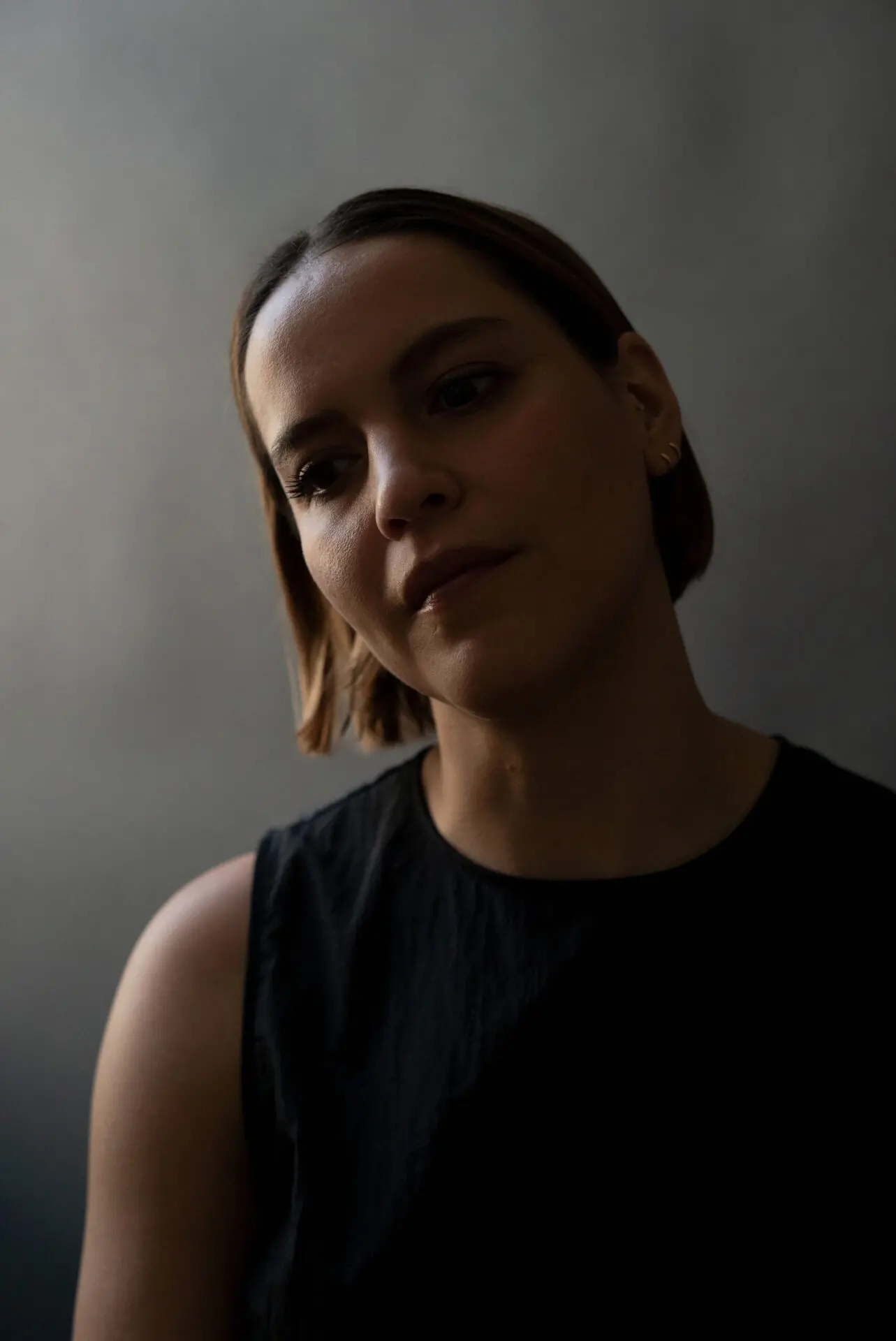 A woman with short brown hair and a sleeveless black top looks slightly downward with a calm, thoughtful expression, as if deep in reflection during an Eloisa Amezcua interview, against a dark, neutral background.