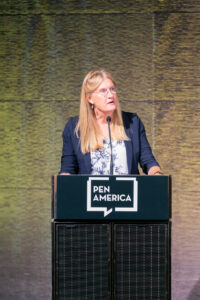 A woman with long blonde hair, wearing glasses and a navy blazer over a white patterned blouse, speaks at a podium with a PEN America sign. The background is textured with shades of yellow and brown.