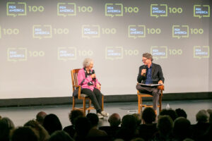 Two people sit on stage in wooden chairs, holding microphones and talking, with a PEN America 100 backdrop behind them. An audience watches from the foreground.