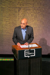 A man in a suit stands at a podium labeled PEN America, speaking into a microphone with papers in front of him against a textured backdrop.