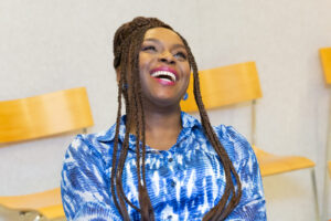 A woman with long braided hair, wearing a blue patterned shirt, smiles and laughs while sitting on a chair. Wooden chairs and a light-colored wall are visible in the background.