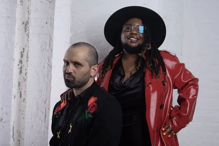 Two people pose in front of a white wall. One pouts in a black jacket with red rose embroidery while the other, channeling Die Jim Crow Records’ bold style, stands behind in a shiny red coat, black dress, sunglasses, and wide-brimmed hat, smiling confidently.