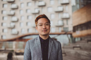 A person with short, light brown hair and glasses, wearing a gray blazer over a dark shirt, stands outdoors in front of a modern building—possibly awaiting a Chen Chen interview.