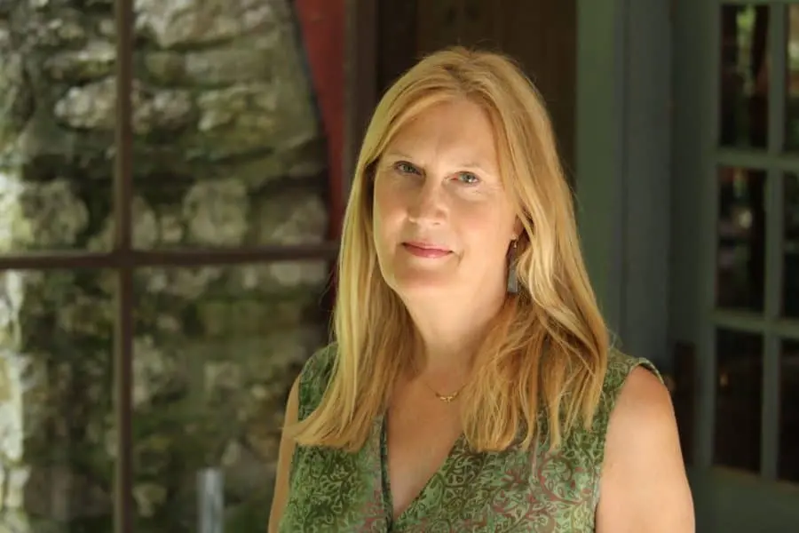 A woman with long blonde hair wearing a green patterned sleeveless top stands indoors near a window at the Pen America 100 Symposium event, looking at the camera with a neutral expression. A stone wall and door are visible in the background.