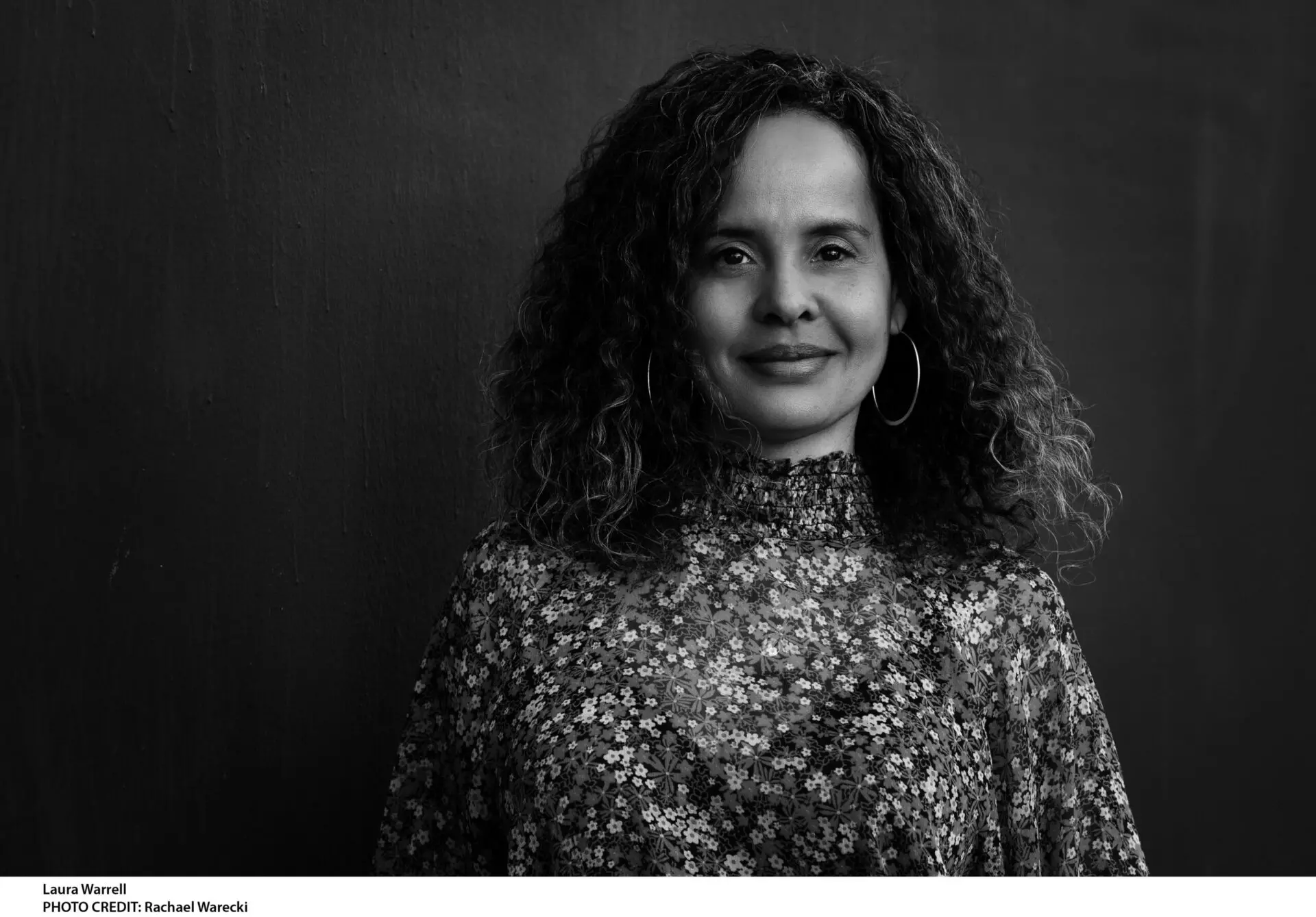 Black and white portrait of a woman with curly hair, wearing hoop earrings and a high-neck, floral-patterned blouse, standing before a plain dark background and smiling softly at the camera—perfect for a Laura Warrell interview feature.