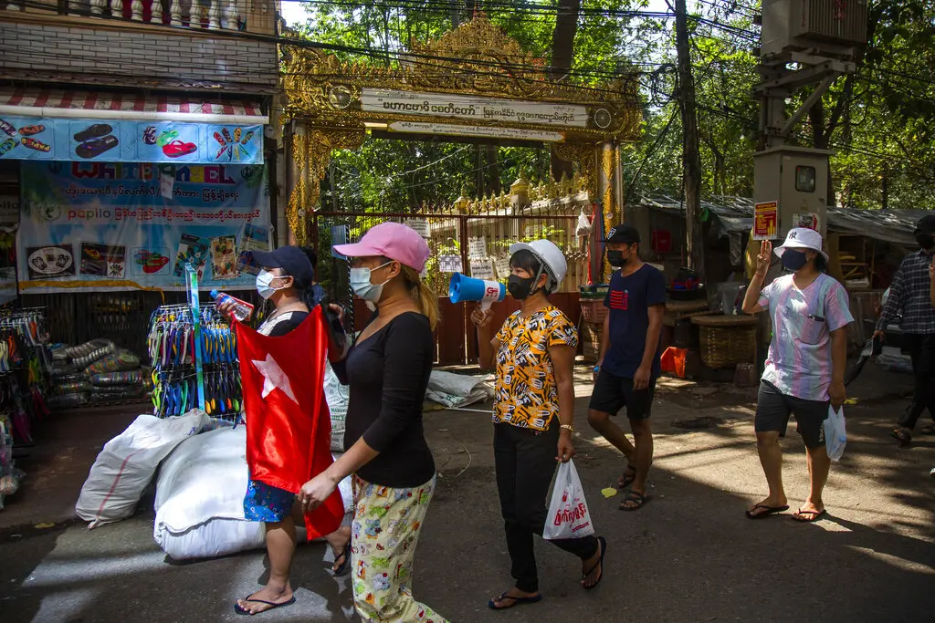 A group of people wearing face masks walk down a street in Myanmar, one person using a megaphone. Clothes and goods are displayed on the left, and a decorated golden gate stands in the background, highlighting resilience amid digital repression.