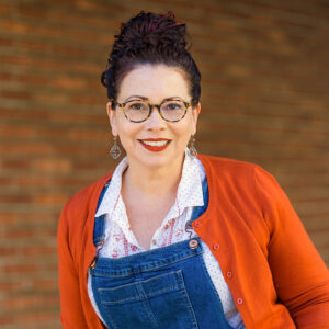 A woman with dark hair in a bun, wearing glasses, red lipstick, earrings, a white patterned shirt, blue denim overalls, and an orange cardigan, smiles in front of a blurred brick wall background during the Donna Barba Higuera interview.