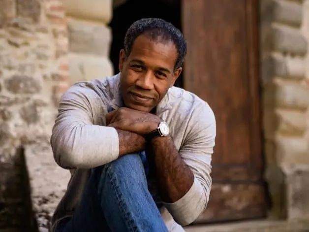Gregory Pardlo, a middle-aged man with short curly hair, sits outdoors, leaning his arms on his knees and smiling gently. He wears a light gray long-sleeve shirt and a watch, with a rustic stone wall and wooden door in the background.