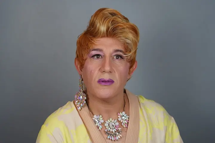A person with short, styled reddish-blonde hair wears purple eyeshadow, pink lipstick, large floral earrings, and a matching necklace—resembling a vibrant look fit for an Iván Monalisa Ojeda interview. They wear a light yellow and cream top against a gray background.