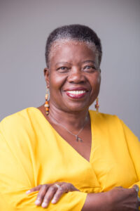 A smiling older woman with short gray hair wearing a bright yellow top and colorful bead earrings stands with arms crossed against a plain background, reminiscent of a Charyl Boyce-Taylor interview.