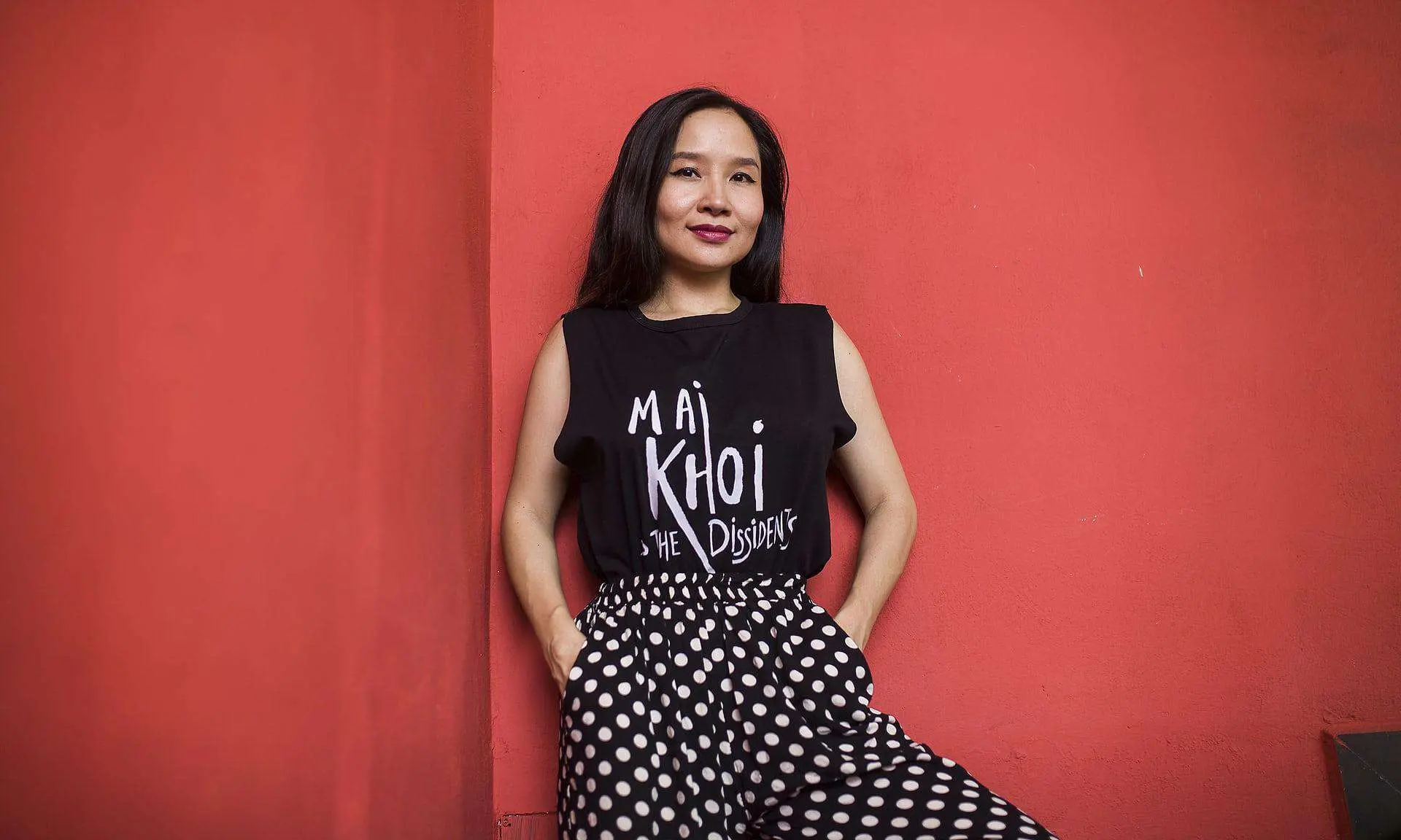 A woman with long dark hair stands against a red wall, wearing a black sleeveless “Mai Khoi & The Dissidents” top—an artist known for advocating freedom of speech in Vietnam—paired with high-waisted black pants with white polka dots. She smiles softly, hands in pockets.