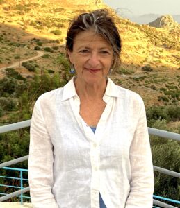 A woman with gray and brown hair, wearing a white shirt over a blue top, stands outdoors on a balcony with hills and greenery behind her, bathed in warm sunlight—like pausing on the map of hope and sorrow.