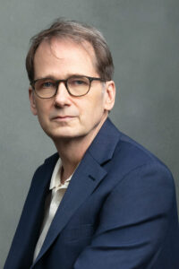 A middle-aged man with short brown hair and glasses wears a navy blue suit and a white shirt, posing against a plain gray background as if ready for a John Weir interview.