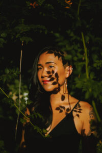 A woman with long dark hair and a tattoo on her arm stands among leafy branches, sunlight casting shadows across her face and body—her contemplative gaze reminiscent of a Cynthia Dewi Oka interview, surrounded by lush greenery.