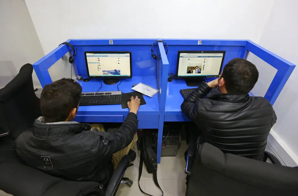 Two people sit at adjacent blue computer cubicles, each using a desktop computer. Both are wearing dark jackets and facing their screens, possibly browsing digital news about Afghanistan or the Taliban in a modern, well-lit setting.