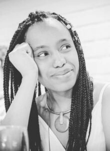 A woman with braided hair rests her head on her hand and smiles thoughtfully during the Rešoketšwe Manenzhe interview. She is wearing a sleeveless top and a chunky necklace. The photo is in black and white.