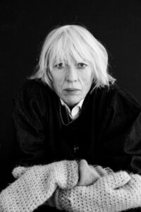 An older woman with pale hair and a serious expression sits against a dark background, wrapped in a chunky knitted blanket—her thoughtful pose reminiscent of a reflective Kristín Ómarsdóttir interview.