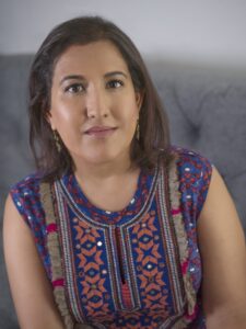 A woman with medium-length brown hair sits on a gray couch, wearing a vibrant blue and orange patterned top with decorative fringe. She looks directly at the camera with a neutral expression, as if ready for a Kavita Das interview.