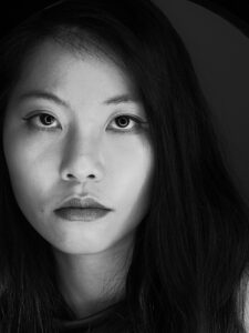 Black and white close-up portrait of a woman with long dark hair, neutral expression, and bold eyeliner, looking directly at the camera—reminiscent of a Kimberly Nguyen interview. Shadows fall on one side of her face.