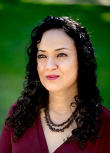 A woman with curly dark hair and a maroon top stands outdoors, wearing a beaded necklace. She looks slightly to the side with a gentle expression, as if deep in thought during an Oindrila Mukherjee interview; blurred green grass is behind her.