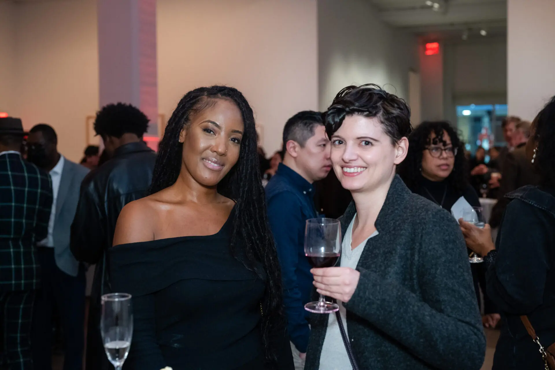Two women pose and smile at an indoor PEN America New Year New Books event. One holds a wine glass as others mingle in the lively and sophisticated atmosphere.