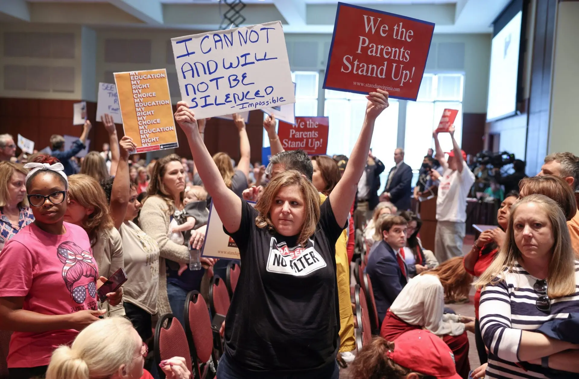 A woman holds a sign reading “I CAN NOT AND WILL NOT BE SILENCED” at a crowded indoor meeting on educational gag orders. Other attendees hold signs saying “We the Parents Stand Up!” expressing protest or advocacy.