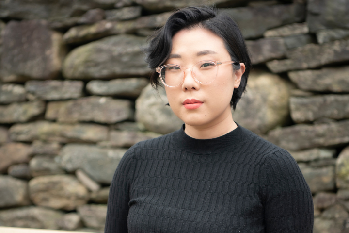 A person with short black hair and glasses, wearing a black ribbed sweater, stands in front of a stone wall, looking calmly at the camera—ready for an interview with Franny Choi.