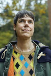 A person with short brown hair wearing a green jacket and a colorful diamond-patterned sweater stands outdoors, looking upward with a slight smile—as if deep in thought after an interview with Isabel Waidner. The background is blurred with trees and autumn colors.
