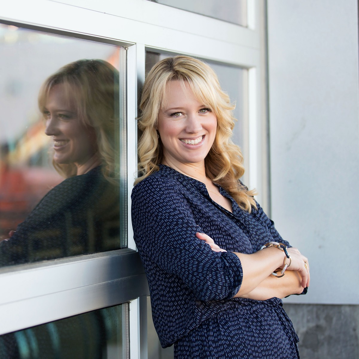 A woman with blonde hair, wearing a dark patterned dress, stands smiling with arms crossed next to a glass window, her reflection visible in the glass—ready to celebrate with her Honeyfund as DeSantis inspires new adventures.