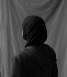 A person wearing a dark hijab and jacket is seen from behind, facing a draped fabric backdrop in this black and white photograph, evoking the reflective mood of an interview with Lamya H. Their face is turned slightly to the side, but remains hidden.