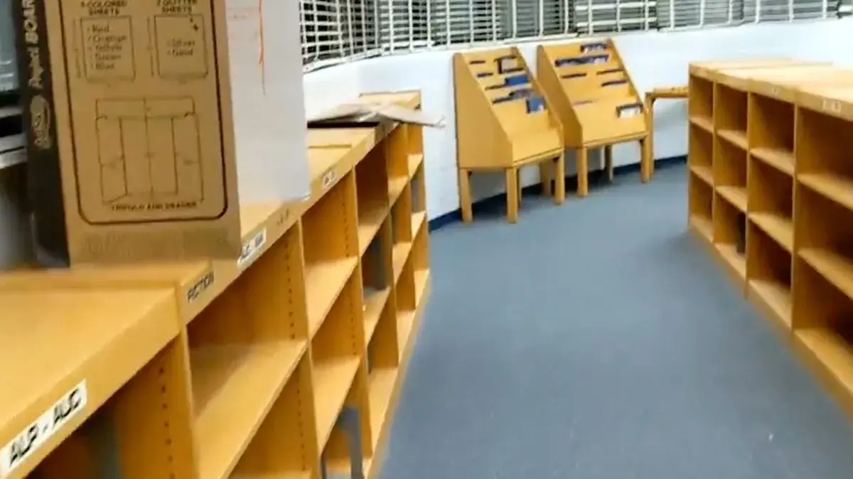 Empty wooden bookshelves and display racks line the blue-carpeted floor of a library, reflecting the impact of Florida book bans. A cardboard box sits on one shelf, while most shelves remain bare, with only a few labels visible.