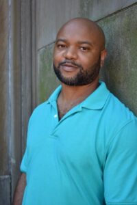 A man with a shaved head and short beard is wearing a teal polo shirt, standing and smiling slightly while leaning against a concrete wall during an interview deShawn Charles Winslow.