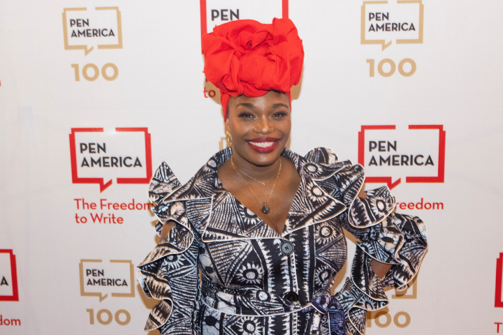 Erika Dickerson-Despenza, wearing a bold patterned dress and a large, bright red headwrap, smiles on a red carpet in front of a PEN America step-and-repeat backdrop.