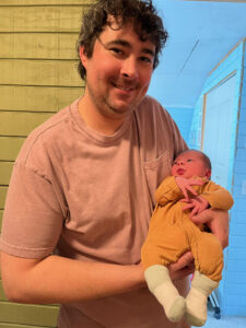 A man with curly brown hair and a pink t-shirt smiles while holding a newborn baby dressed in yellow and white socks, standing indoors near a green wooden wall, reminiscent of a tender scene in a Morgan Talty novel.